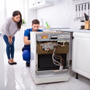 Smiling Woman Behind Technician Repairing Dishwasher In Kitchen